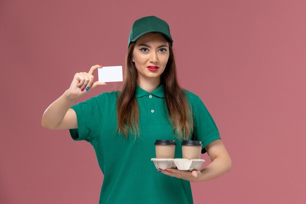 Mensajero femenino de vista frontal en uniforme verde y capa sosteniendo tazas de café de entrega y tarjeta en la entrega uniforme de trabajo de servicio de pared rosa