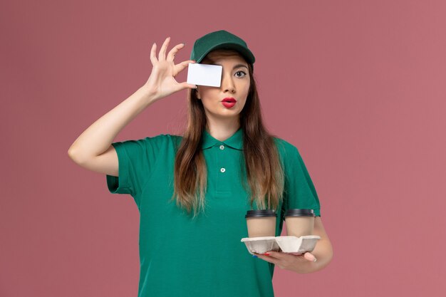 Mensajero femenino de vista frontal en uniforme verde y capa sosteniendo tazas de café de entrega y tarjeta en la entrega uniforme de trabajo de servicio de pared rosa