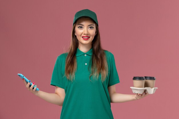 Mensajero femenino de vista frontal en uniforme verde y capa sosteniendo tazas de café de entrega y teléfono en la entrega uniforme de servicio de pared rosa