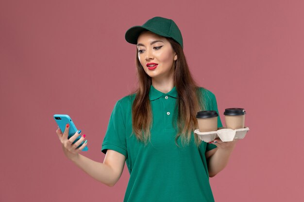 Mensajero femenino de vista frontal en uniforme verde y capa sosteniendo tazas de café de entrega y teléfono en la entrega uniforme de trabajo de servicio de pared rosa