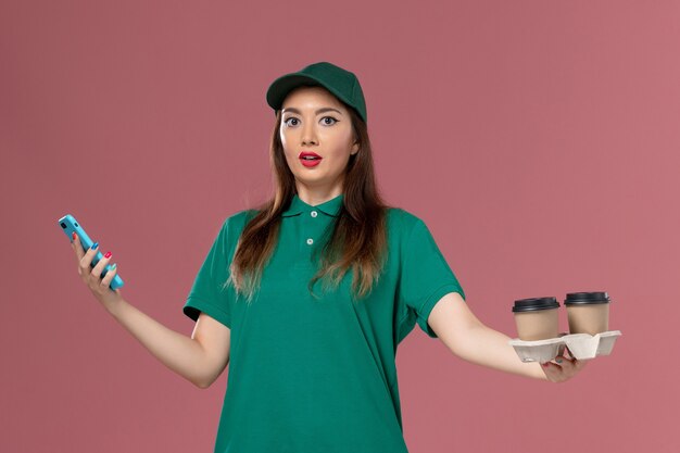 Mensajero femenino de vista frontal en uniforme verde y capa sosteniendo tazas de café de entrega y teléfono en servicio de escritorio rosa entrega uniforme de trabajo