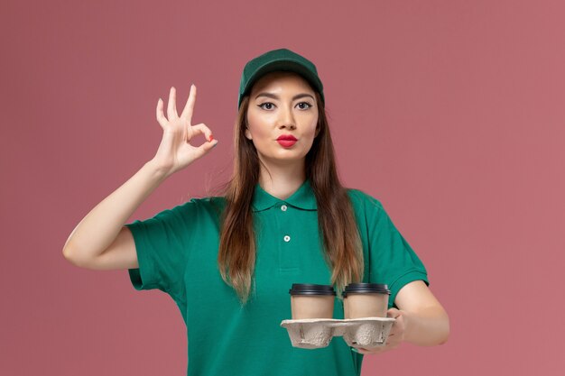 Mensajero femenino de vista frontal en uniforme verde y capa sosteniendo tazas de café de entrega en el trabajador de trabajo de entrega uniforme de servicio de pared rosa