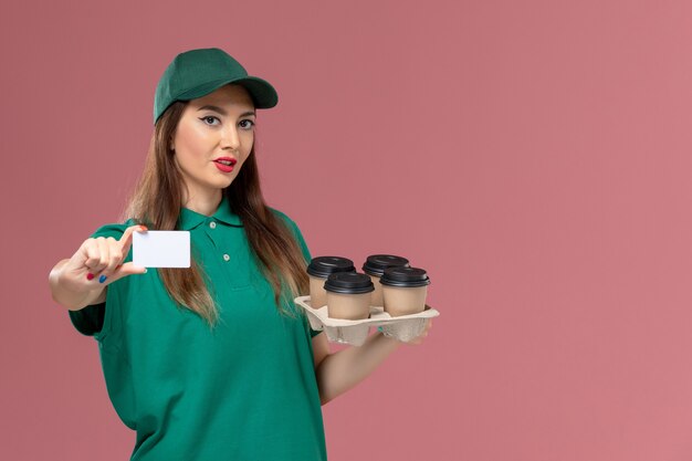 Mensajero femenino de vista frontal en uniforme verde y capa con tarjeta y tazas de café de entrega en servicio de escritorio rosa entrega uniforme