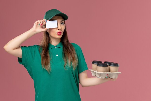 Mensajero femenino de vista frontal en uniforme verde y capa con tarjeta y tazas de café de entrega en el trabajador de trabajo de entrega uniforme de servicio de pared rosa