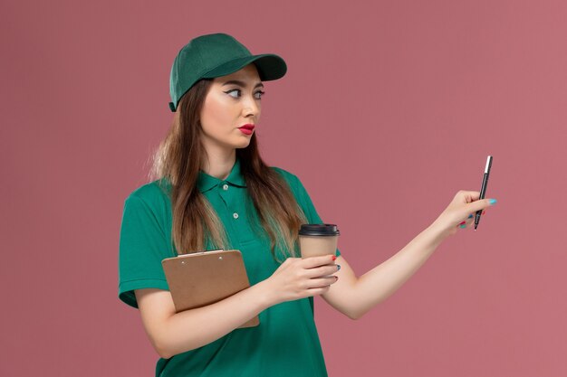 Mensajero femenino de vista frontal en uniforme verde y capa con taza de café de entrega y bolígrafo de bloc de notas en servicio de empresa de escritorio rosa entrega uniforme