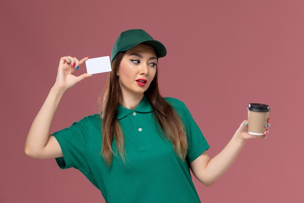 Mensajero femenino de vista frontal en uniforme verde y capa con taza de café de entrega con tarjeta en el trabajo de entrega uniforme de trabajo de servicio de escritorio rosa