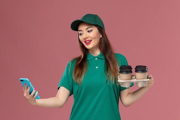 Mensajero femenino de vista frontal en uniforme verde y capa usando teléfono y sosteniendo tazas de café de entrega en el trabajo de entrega uniforme de servicio de pared rosa