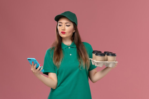 Mensajero femenino de vista frontal en uniforme verde y capa usando teléfono y sosteniendo tazas de café de entrega en el trabajo de entrega uniforme de servicio de pared rosa