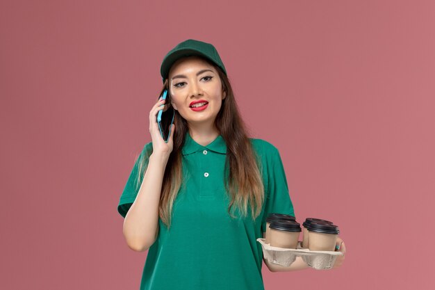 Mensajero femenino de vista frontal en uniforme verde hablando por teléfono y sosteniendo tazas de café de entrega en el trabajo de entrega uniforme de servicio de pared rosa