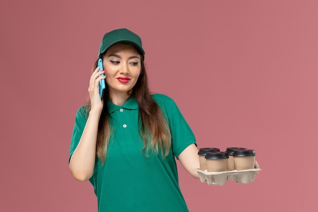 Mensajero femenino de vista frontal en uniforme verde hablando por teléfono y sosteniendo tazas de café de entrega en el trabajo de entrega uniforme de servicio de piso rosa