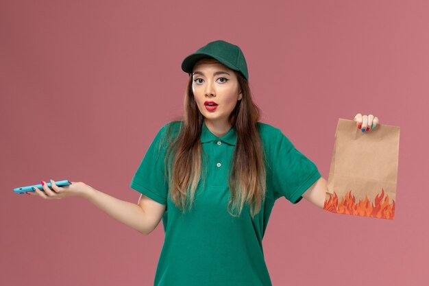 Mensajero femenino de vista frontal en uniforme verde que usa el teléfono y que sostiene el paquete de alimentos en el trabajador de trabajo de entrega uniforme de servicio de pared rosa