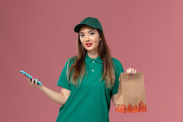 Mensajero femenino de vista frontal en uniforme verde que usa el teléfono y que sostiene el paquete de alimentos en el trabajador de trabajo de entrega uniforme de servicio de pared rosa