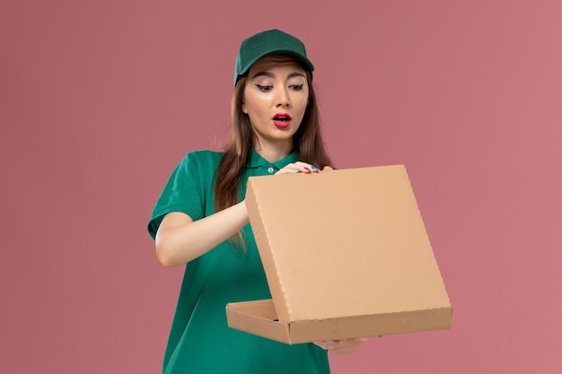 Mensajero femenino de vista frontal en uniforme verde sosteniendo y abriendo la caja de entrega de alimentos en la empresa de repartidor uniforme de servicio de pared rosa