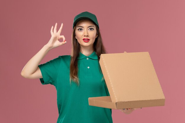 Mensajero femenino de vista frontal en uniforme verde sosteniendo una caja de entrega de alimentos vacía en la empresa de chica de entrega uniforme de servicio de pared rosa