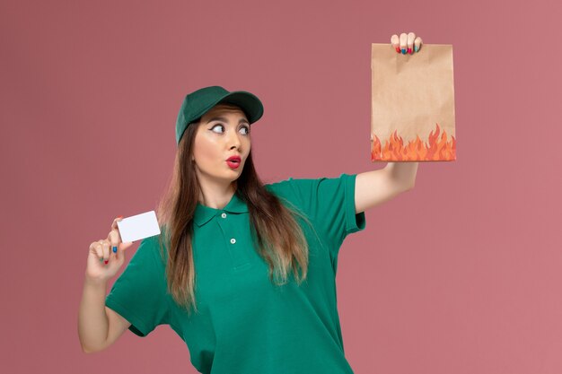 Mensajero femenino de vista frontal en uniforme verde con tarjeta blanca y paquete de alimentos en trabajo de entrega de trabajador uniforme de servicio de pared rosa