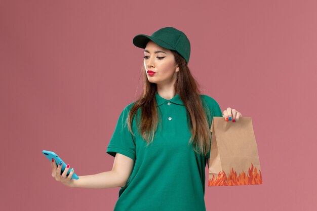 Mensajero femenino de vista frontal en uniforme verde usando el teléfono y sosteniendo el paquete de alimentos en el trabajo de entrega uniforme de servicio de pared rosa