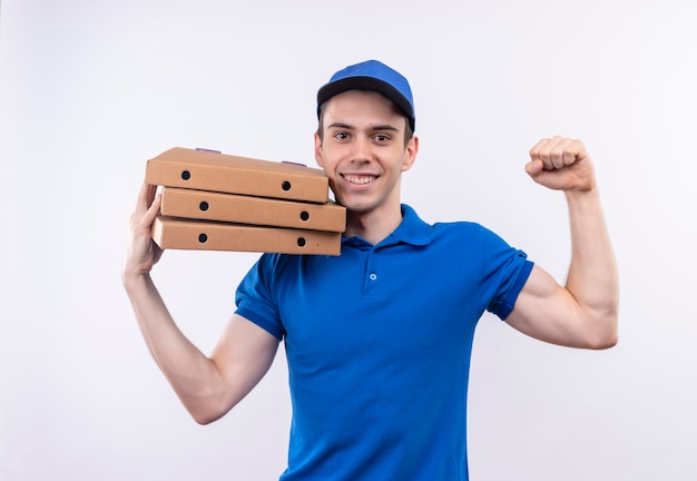 Mensajero joven con uniforme azul y gorra azul haciendo feliz puño y sosteniendo cajas