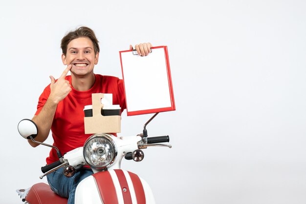 Mensajero joven en uniforme rojo sentado en scooter con pedidos y documentos haciendo gesto de sonrisa en la pared amarilla