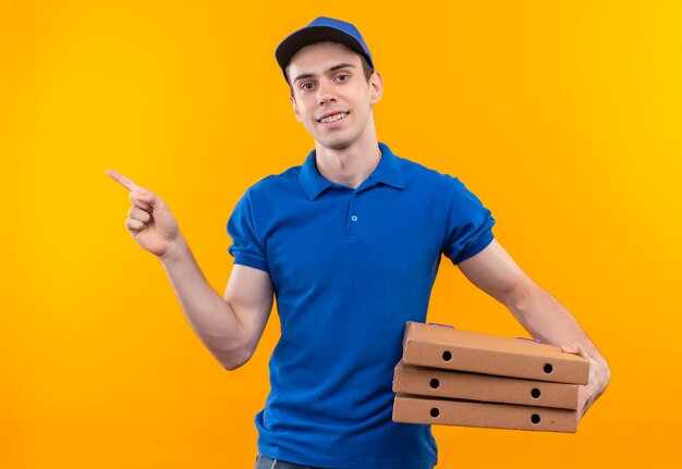 Mensajero joven vestido con uniforme azul y gorra azul haciendo feliz dedo índice a la derecha contiene cajas