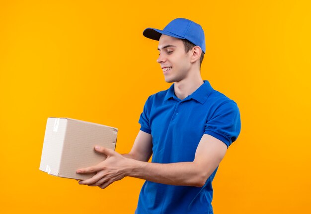 Mensajero joven vistiendo uniforme azul y gorra azul felizmente sostiene una caja