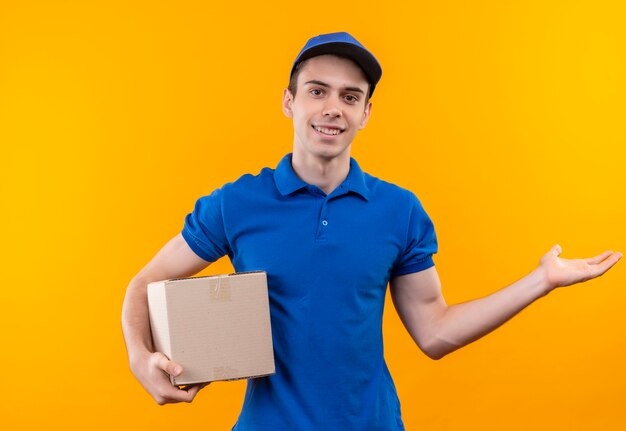 Mensajero joven vistiendo uniforme azul y gorra azul felizmente sostiene una caja