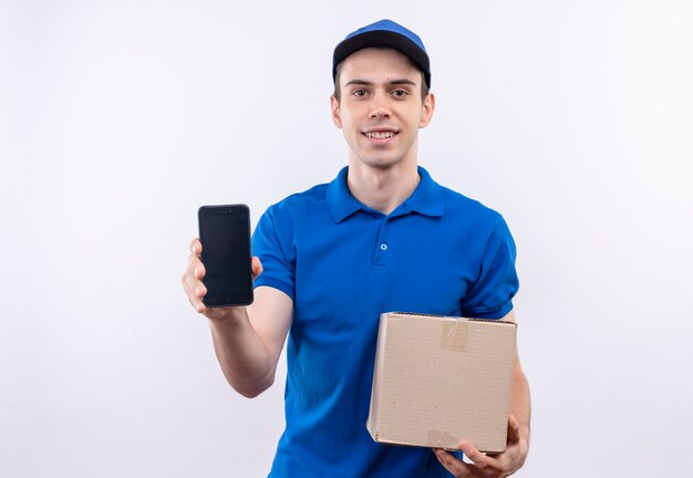 Mensajero joven vistiendo uniforme azul y gorra azul felizmente sostiene un teléfono y una bolsa