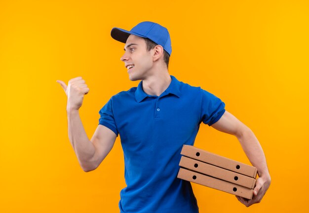 Mensajero joven vistiendo uniforme azul y gorra azul haciendo felices pulgares a la derecha tiene cajas