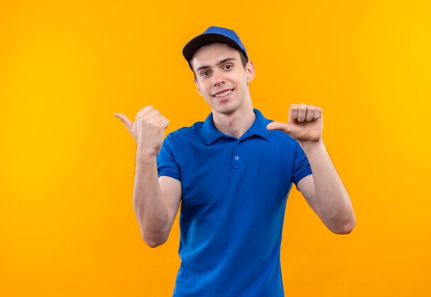 Mensajero joven vistiendo uniforme azul y gorra azul haciendo felices pulgares a la izquierda