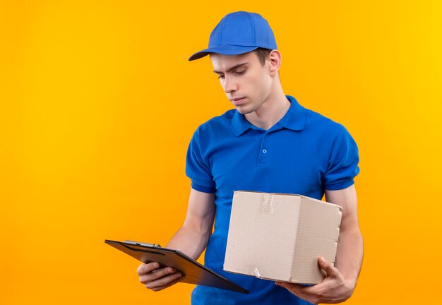 Mensajero joven vistiendo uniforme azul y gorra azul mira en el portapapeles y sostiene una caja