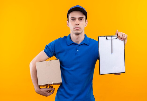 Mensajero joven vistiendo uniforme azul y gorra azul sostiene seriamente una caja y portapapeles