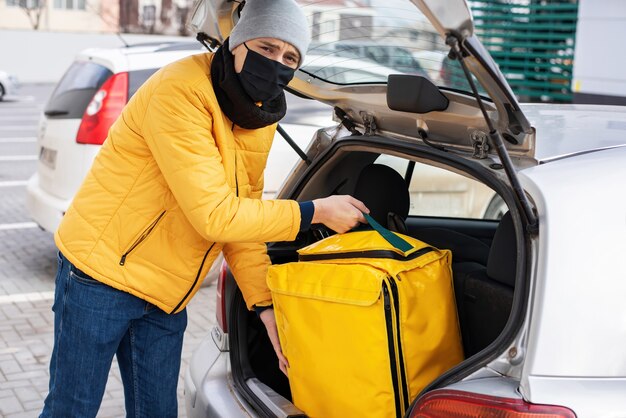 Mensajero con máscara médica negra sacando mochila amarilla del coche. Servicio de comida a domicilio