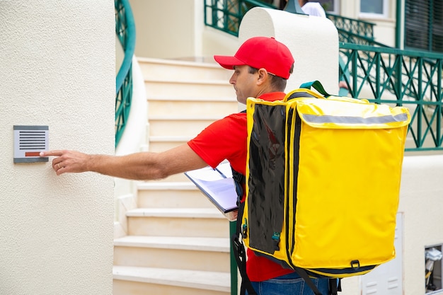 Mensajero masculino con bolsa térmica y timbre de timbre portapapeles. Repartidor serio en uniforme rojo empujando el timbre de la puerta, de pie al aire libre y entregando el pedido Servicio de entrega a domicilio y concepto de publicación