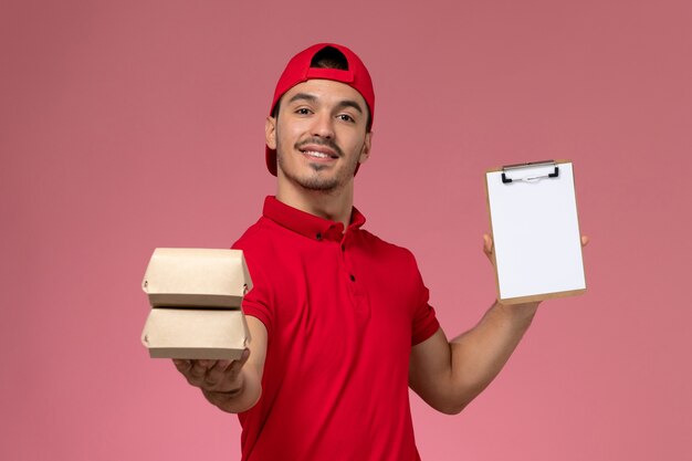 Mensajero masculino joven de la vista frontal en capa uniforme roja que sostiene el bloc de notas y el pequeño paquete con comida sobre fondo rosa.