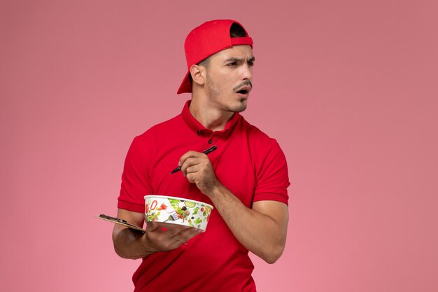Mensajero masculino joven de la vista frontal en capa uniforme roja que sostiene el cuenco de la entrega con el bloc de notas y el bolígrafo sobre fondo rosa.