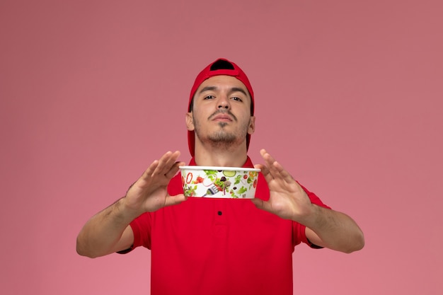 Foto gratuita mensajero masculino joven de la vista frontal en capa uniforme roja que sostiene el cuenco de la entrega en el fondo rosado claro.