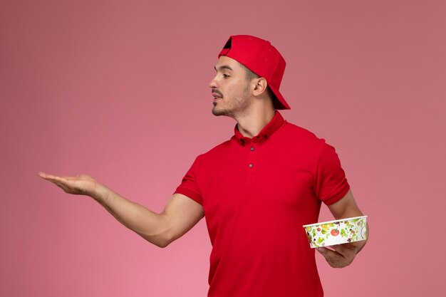 Mensajero masculino joven de la vista frontal en capa uniforme roja que sostiene el cuenco de la entrega en el fondo rosado claro.