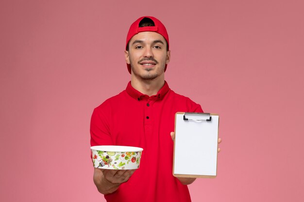 Mensajero masculino joven de la vista frontal en capa uniforme roja que sostiene el cuenco redondo de la entrega y el bloc de notas sonriendo sobre fondo rosa.