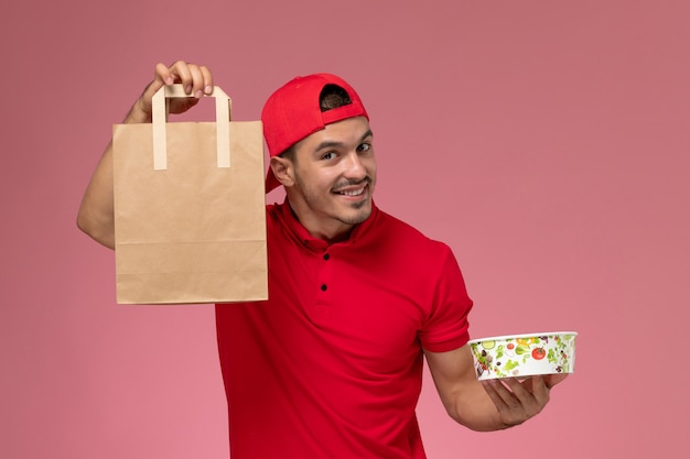 Foto gratuita mensajero masculino joven de la vista frontal en capa uniforme roja que sostiene el paquete de alimentos y el tazón con una sonrisa en el fondo rosa.