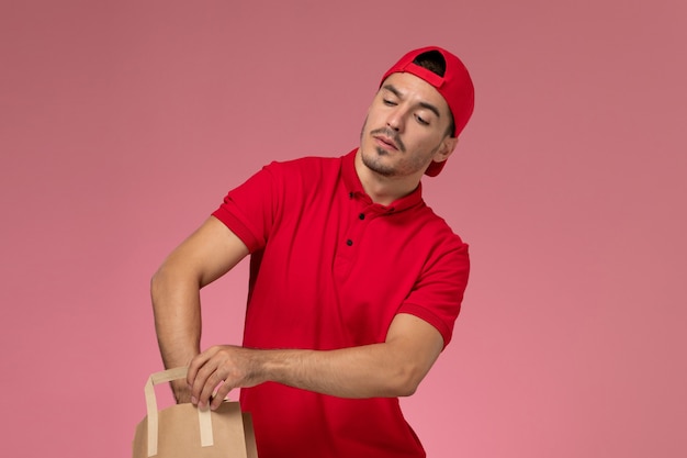 Foto gratuita mensajero masculino joven de la vista frontal en capa uniforme roja que sostiene el paquete de papel del alimento en el fondo rosado.