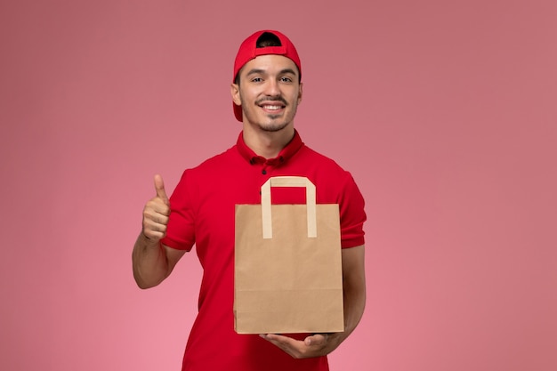 Mensajero masculino joven de la vista frontal en capa uniforme roja que sostiene el paquete de papel del alimento en el fondo rosado.