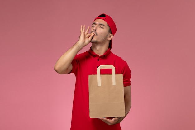 Mensajero masculino joven de la vista frontal en capa uniforme roja que sostiene el paquete de papel del alimento en el fondo rosado.