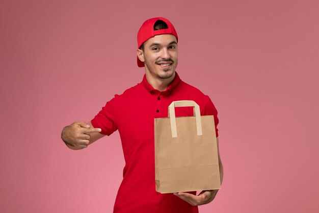 Mensajero masculino joven de la vista frontal en capa uniforme roja que sostiene el paquete de papel del alimento en el fondo rosado.