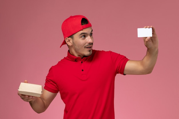 Foto gratuita mensajero masculino joven de la vista frontal en capa uniforme roja que sostiene el paquete pequeño de la entrega con la tarjeta blanca en el fondo rosado.