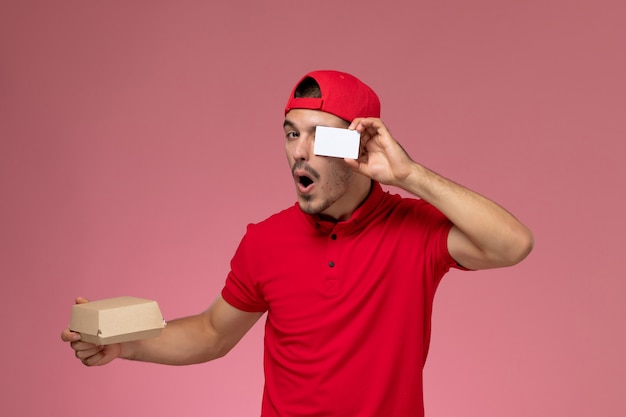 Mensajero masculino joven de la vista frontal en capa uniforme roja que sostiene el paquete pequeño de la entrega con la tarjeta blanca en el fondo rosado.