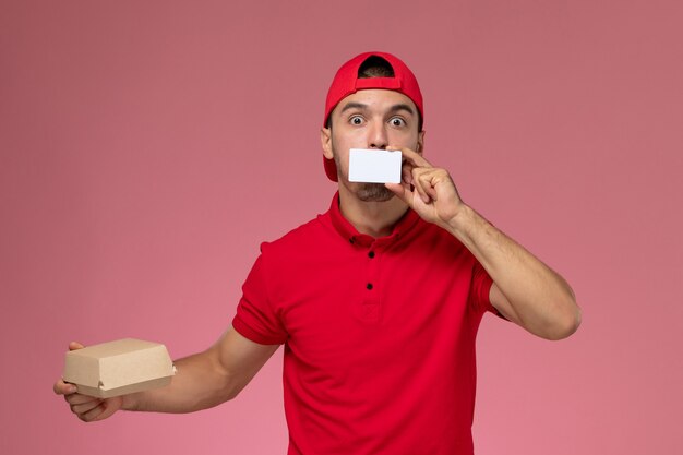 Mensajero masculino joven de la vista frontal en capa uniforme roja que sostiene el pequeño paquete de entrega con tarjeta blanca sobre fondo rosa claro.