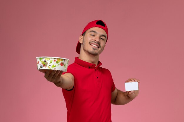 Mensajero masculino joven de la vista frontal en capa uniforme roja que sostiene la tarjeta plástica blanca y el cuenco de la entrega en el fondo rosa claro.