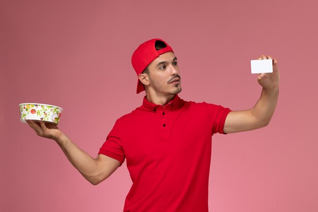 Mensajero masculino joven de la vista frontal en capa uniforme roja que sostiene la tarjeta plástica blanca y el cuenco de la entrega en el fondo rosa claro.