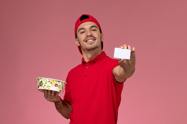 Mensajero masculino joven de la vista frontal en capa uniforme roja que sostiene la tarjeta plástica blanca y el cuenco de la entrega en el fondo rosa claro.