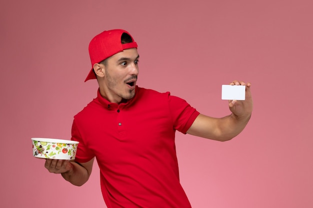 Mensajero masculino joven de la vista frontal en capa uniforme roja que sostiene la tarjeta plástica blanca y el cuenco de la entrega en el fondo rosa claro.