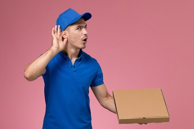 Mensajero masculino en uniforme azul con caja de entrega de comida tratando de escuchar en rosa, servicio de entrega uniforme de trabajador de trabajo